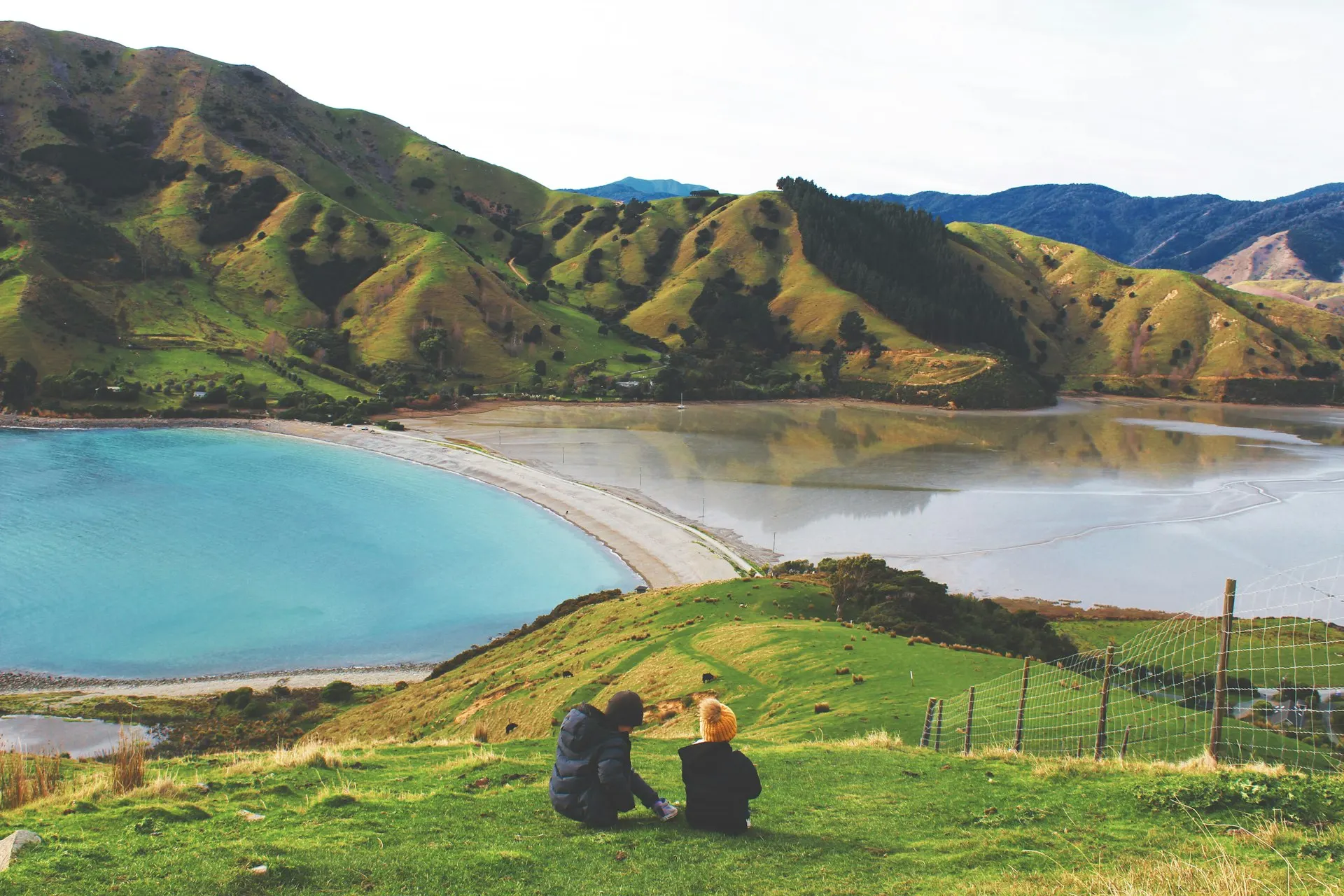 A couple looking out over a turquoise New Zealand bay with their camping gear